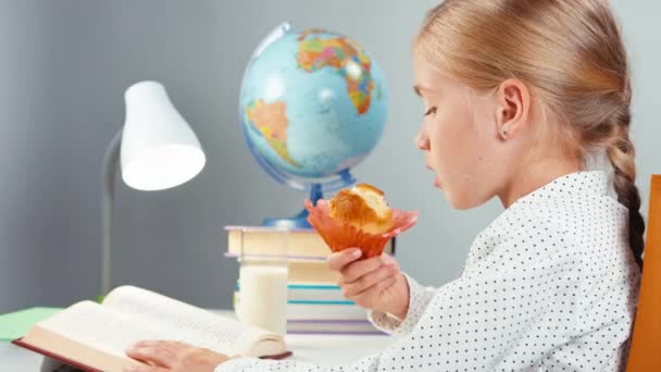 Közelkép iskolás portré 7-8 év enni muffin fogyasztói tej és könyvet olvas, és ül a pult fehér háttér — Stock videók