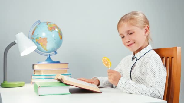 Schoolmeisje lezen van een boek op het Bureau en likken lollipop — Stockvideo