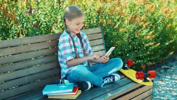 Gelukkig meisje typen sms op haar smartphone zittend op het bankje in het park en glimlachend in de camera. Bovenaanzicht — Stockvideo