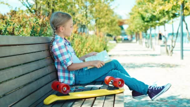 Retrato chica 7-8 años leyendo libro y sentado en el banco en el parque y sonriendo a la cámara — Vídeo de stock