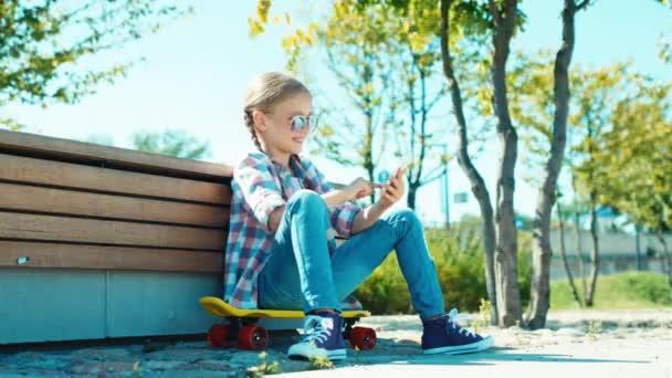 Ragazza felice 7-8 anni in occhiali da sole giocare nel gioco sul suo smartphone seduto sullo skateboard vicino alla panchina — Video Stock