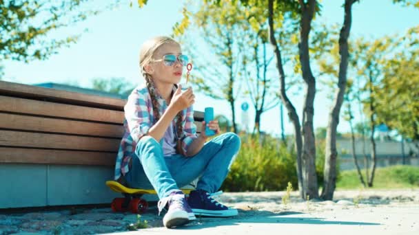 Portret kind meisje 7-8 jaar bellen blazen en zittend op het skateboard in de buurt van de Bank in het park — Stockvideo