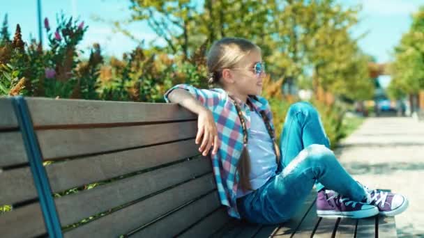 Девушка 7-8 лет в солнцезащитных очках сидит на скамейке и улыбается на камеру в парке — стоковое видео