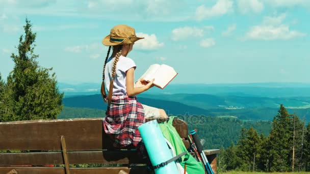 Little hiker girl child 7-8 years old reading book and sitting on the bench on mountains background. Zooming — Stock Footage Little hiker girl child 7-8 years old reading book and sitting on the bench on mountains background. Zooming Stock Video