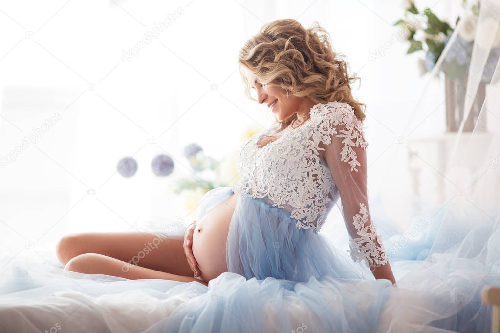 Pretty smiling pregnant girl is wearing blue boudoir dress lying in bed