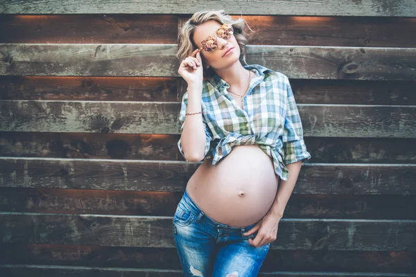 Красивая улыбающаяся беременная девушка носит повседневную одежду и солнцезащитные очки позирует над деревянной стеной — стоковое фото