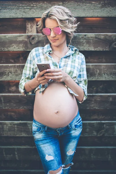 Frumoasă fată gravidă zâmbitoare poartă haine casual și ochelari de soare se prezintă peste perete de lemn — Fotografie, imagine de stoc