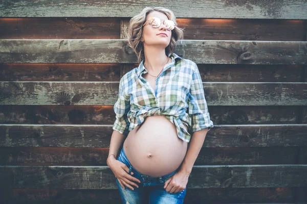 Frumoasă fată gravidă zâmbitoare poartă haine casual și ochelari de soare se prezintă peste perete de lemn — Fotografie, imagine de stoc