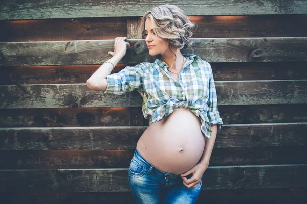 Belle fille enceinte souriante porte des vêtements décontractés pose sur un mur en bois — Photo