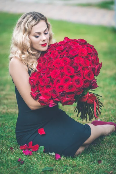 Elegancka blond kobieta siedzi na zielony trawnik z wielki bukiet czerwonych róż 101 — Zdjęcie stockowe