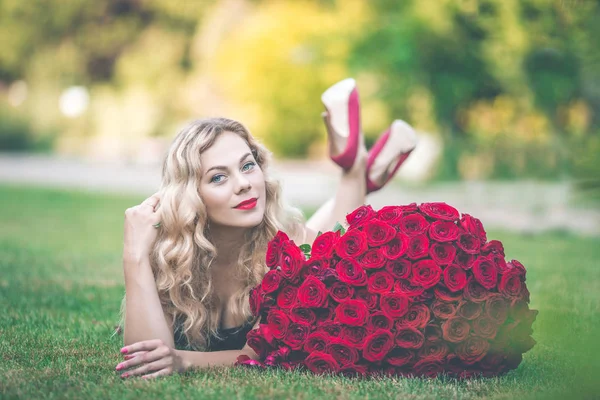 优雅美丽的金发女郎穿着黑色的时尚礼服在绿色的草坪上, 大花束101红玫瑰 — 图库照片