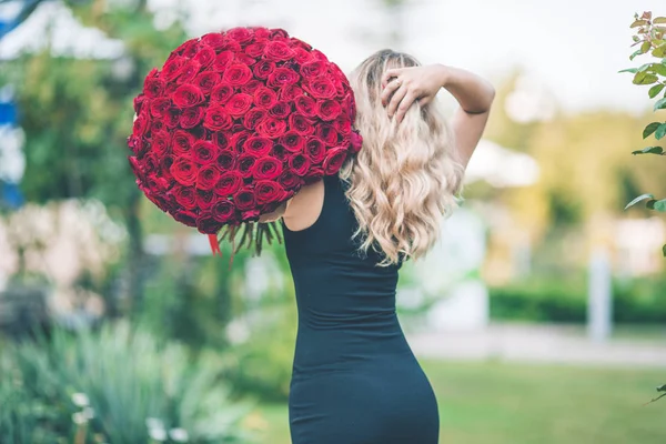 Vista posterior de la mujer hermosa elegante está usando vestido de moda negro en el césped verde con gran ramo de 101 rosas rojas — Foto de Stock