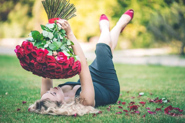 Elegant beautiful blonde woman is wearing black fashion dress lying on green lawn holding with big bouquet of 101 red roses — Stock Photo, Image