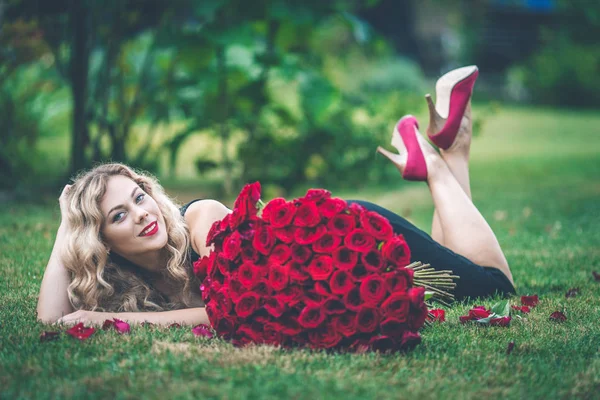 Elegant vacker blond kvinna bär svart mode klä liggande på grön gräsmatta håller med stor bukett 101 röda rosor — Stockfoto