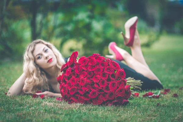 优雅美丽的金发女郎穿着黑色的时尚礼服躺在绿色的草坪上, 捧着101朵红玫瑰的大花束 — 图库照片