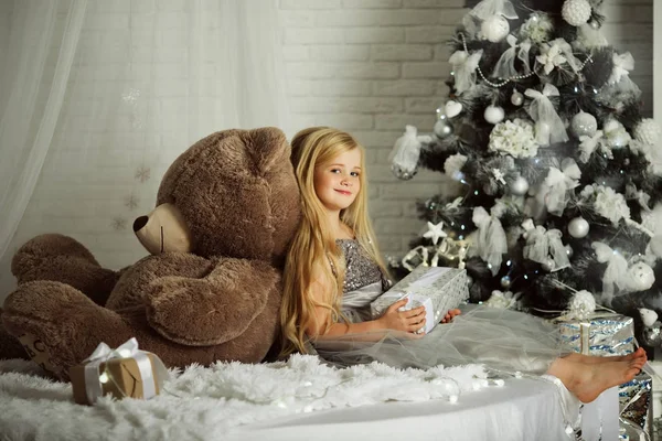 Feliz Navidad y Felices Fiestas. Bonita chica rubia con el pelo largo está sosteniendo gran osito de peluche —  Fotos de Stock