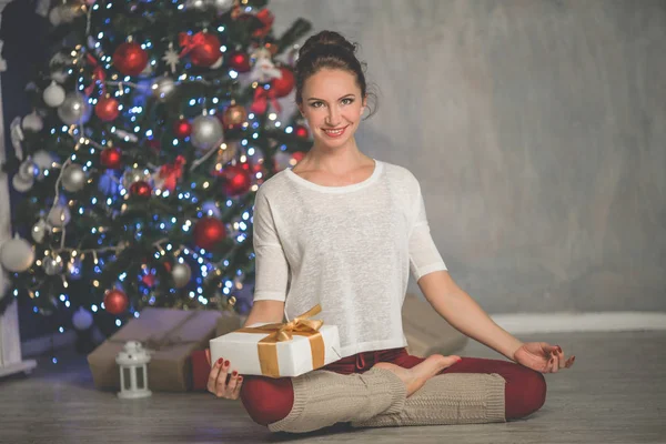Hermosa mujer sonriente flexible con caja de regalo está haciendo ejercicio de estiramiento cerca del árbol de Navidad de moda decorado en casa, deportes y concepto de vacaciones —  Fotos de Stock