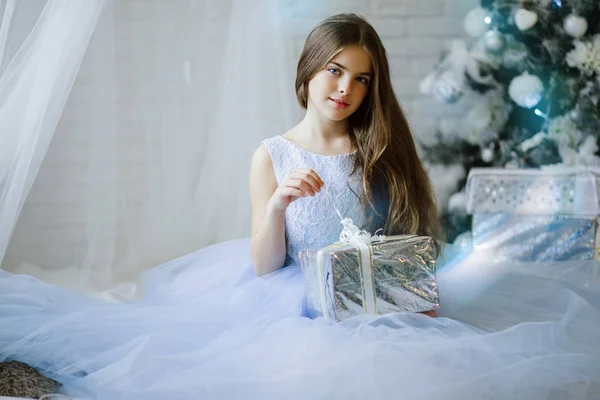 Krásná dívka sedí poblíž vánoční stromeček s stříbrná dárková krabička — Stock fotografie