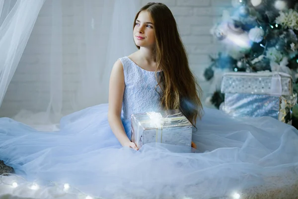 Krásná dívka sedí poblíž nazdobený vánoční stromeček s stříbrná dárková krabička — Stock fotografie