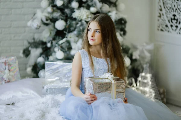 Beautiful girl is sitting near decorated christmas tree with silver gift box — Stock Photo, Image