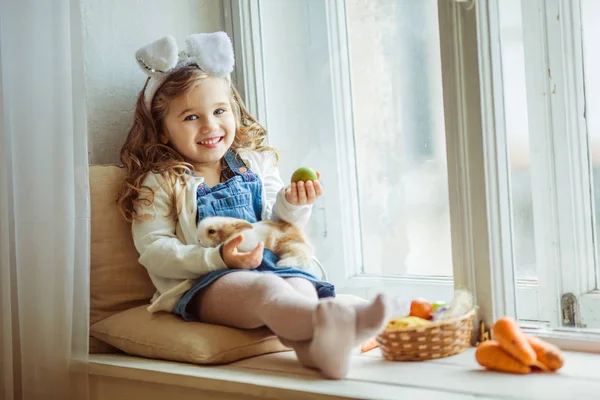 Cute happy little child girl is wearing bunny ears on Easter day holding her friend little colorful rabbit — Stock Photo, Image