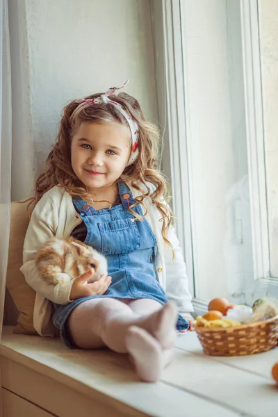 Ziemlich glückliches Kind Mädchen sitzt auf Fensterbank mit ihrem Freund kleine bunte Kaninchen, Osterferienkonzept — Stockfoto