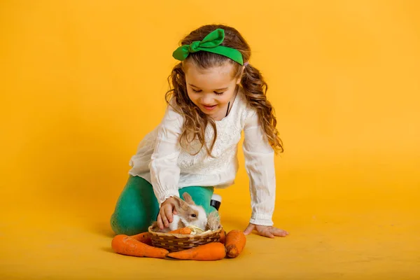 Docela šťastné dítě dívka s mrkví a její přítel králík málo barevné, Velikonoční svátek koncept izolovaných na žluté — Stock fotografie