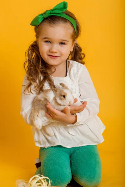 Pretty happy child girl is holding her friend little colorful rabbit, Easter holiday concept isolated on yellow — Stock Photo, Image