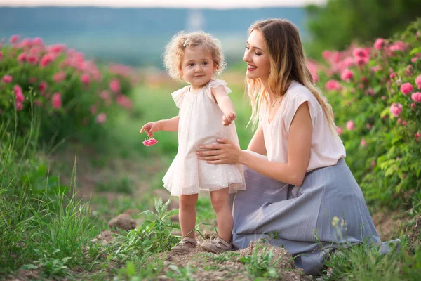 Jovem mãe com sua filha estão andando no jardim da primavera com flores de rosas rosa, hora do pôr do sol — Fotografia de Stock