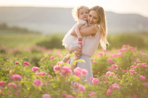 Young mother with her pretty curly daughter are walking in spring garden with pink blossom roses flowers, sunset time — Stock Photo, Image