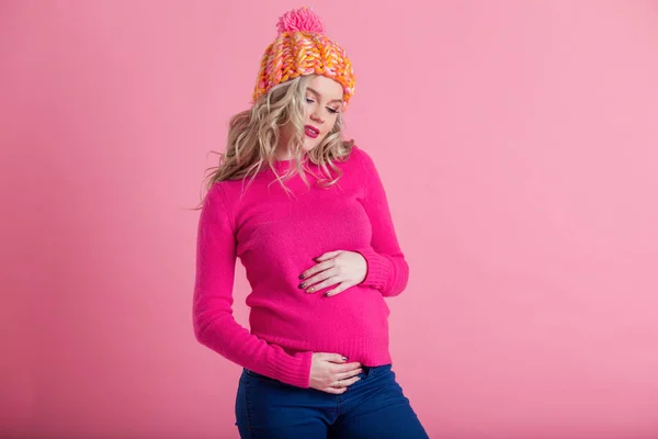 Blonde happy pregnant woman is wearing warm winter clothes hat and sweater isolated on pink background — Stock Photo, Image