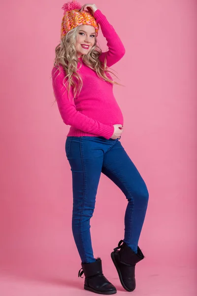 Rubia mujer embarazada feliz está usando ropa de invierno cálido sombrero y suéter aislado sobre fondo rosa — Foto de Stock