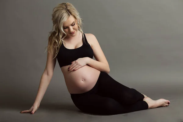 Incinta bella donna bionda indossa vestiti neri sportivi facendo esercizi di yoga isolati su grigio, concetto di vita sana — Foto Stock