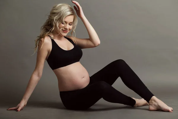 Mujer rubia hermosa embarazada está usando ropa negra deportiva haciendo ejercicios de yoga aislados en gris, concepto de vida saludable — Foto de Stock