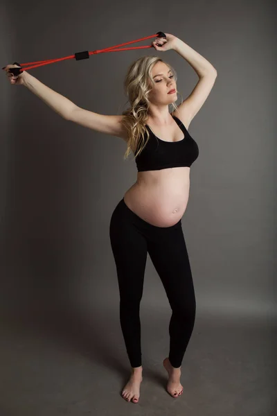 Pregnant beautiful blonde woman is wearing sportive black clothes doing exercises with rubber band isolated on gray, healthy life concept — Stock Photo, Image