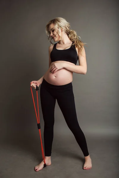 Embarazada hermosa mujer rubia está usando ropa deportiva negro haciendo ejercicios con goma banda aislada en gris, concepto de vida saludable — Foto de Stock