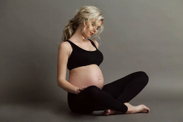 Mujer rubia hermosa embarazada está usando ropa negra deportiva haciendo ejercicios de yoga aislados en gris, concepto de vida saludable —  Fotos de Stock