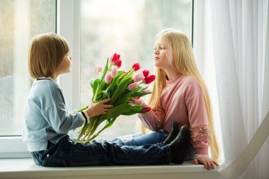 Çok güzel çocuklar mutlu küçük çocuk bir sevimli kız buket Pembe Lale çiçek veren pencere pervazına üzerinde oturuyor. Paskalya kavramı