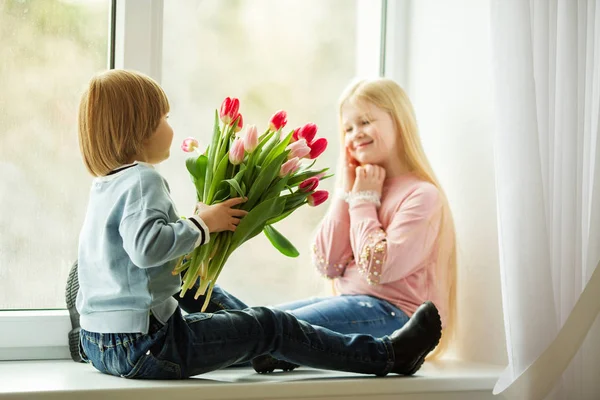 Crianças bonitas estão sentadas no peitoril da janela, menino feliz dando um buquê bonito menina de flores de tulipa rosa. Conceito de Páscoa — Fotografia de Stock