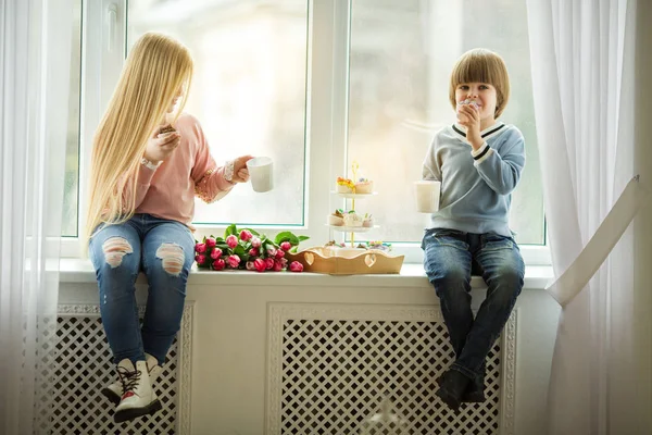 As crianças bonitas estão sentadas no peitoril da janela e comendo bolos com chá, menino e menina bonito com buquê de flores de tulipa rosa. Conceito de Páscoa — Fotografia de Stock