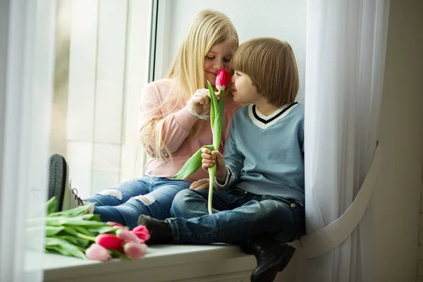 As crianças bonitas estão sentadas no peitoril da janela, menino pequeno feliz e menina bonito com buquê de flores de tulipa rosa. Conceito de Páscoa — Fotografia de Stock