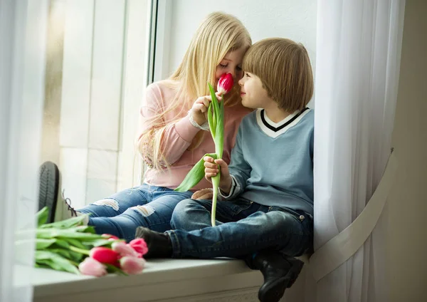 As crianças bonitas estão sentadas no peitoril da janela, menino pequeno feliz e menina bonito com buquê de flores de tulipa rosa. Conceito de Páscoa — Fotografia de Stock