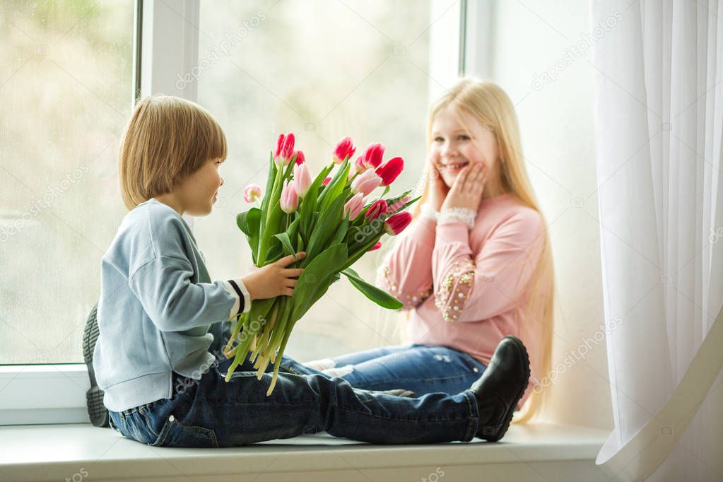 Pretty kids are sitting on window sill , happy little boy giving a cute girl bouquet of pink tulip flowers. Easter concept