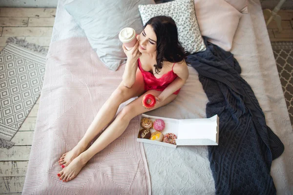 젊은 여자는 초콜릿 도넛의 상자와 침대에서 커피 한잔으로 침대에 누워 — 스톡 사진