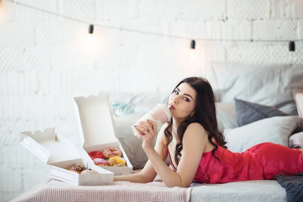 Bella ragazza è sdraiata a letto e bere caffè con ciambelle, ora di colazione — Foto Stock