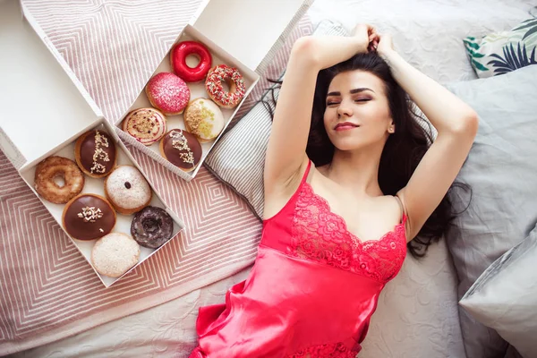 Menina morena bonita está deitada na cama com donuts doces, tempo de café da manhã, conceito de bom dia — Fotografia de Stock
