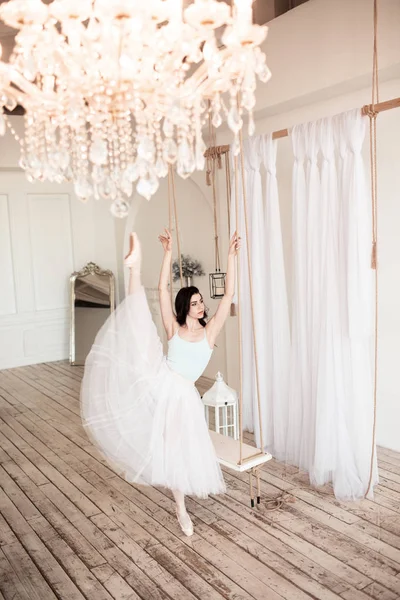 Young pretty ballerina is wearing white skirt dancing near swing in studio with light interior and wooden floor — Stock Photo, Image