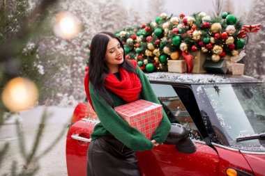 Güzel genç kız kırmızı arabanın yanında elinde Noel ağacı süslenmiş bir hediye kutusu tutuyor. Tatil konsepti..