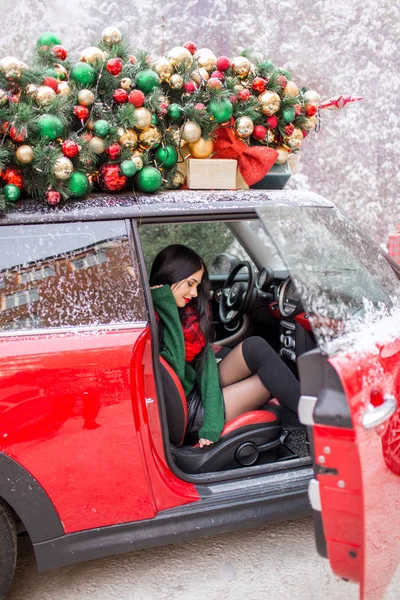 Hübsches junges Mädchen sitzt in rotem Auto mit geschmücktem Weihnachtsbaum auf dem Dach. — Stockfoto