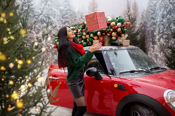 Красива молода дівчина тримає подарункову коробку біля червоного автомобіля з прикрашеною ялинкою на даху, концепція свята . — стокове фото